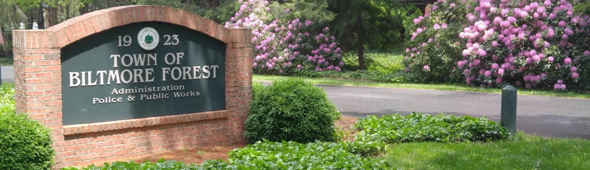 City entrance sign for the Town of Biltmore Forest, established in 1923, featuring administration, police, and public works departments, with pink flowers and greenery in the background.